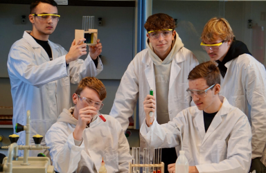 Eine Schülergruppe in weißen Kitteln und mit Schutzbrillen bei einem Experiment im naturwissenschaftlichen Unterricht