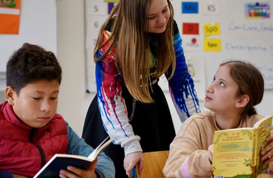 Ein Schüler liest in einem Buch, daneben unterhalten sich zwei Schülerinnen