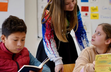Ein Schüler liest in einem Buch, daneben unterhalten sich zwei Schülerinnen