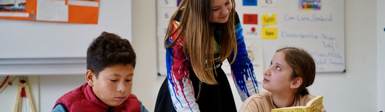 Ein Schüler liest in einem Buch, daneben unterhalten sich zwei Schülerinnen