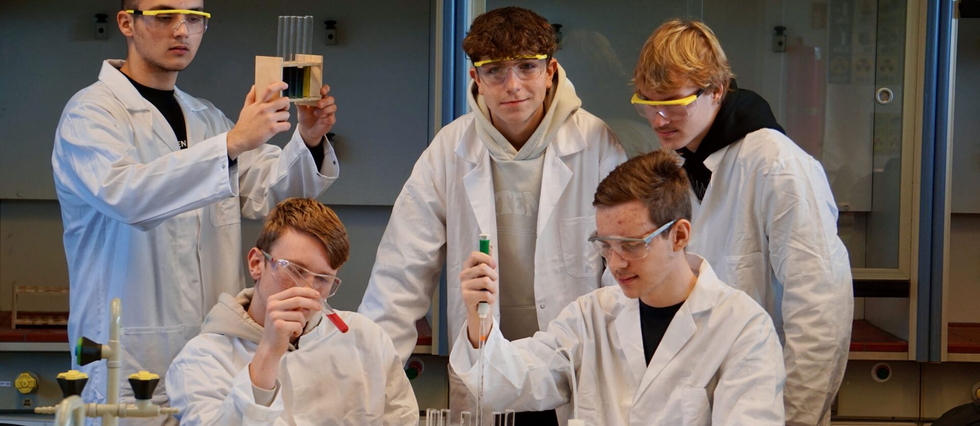 Eine Schülergruppe in weißen Kitteln und mit Schutzbrillen bei einem Experiment im naturwissenschaftlichen Unterricht